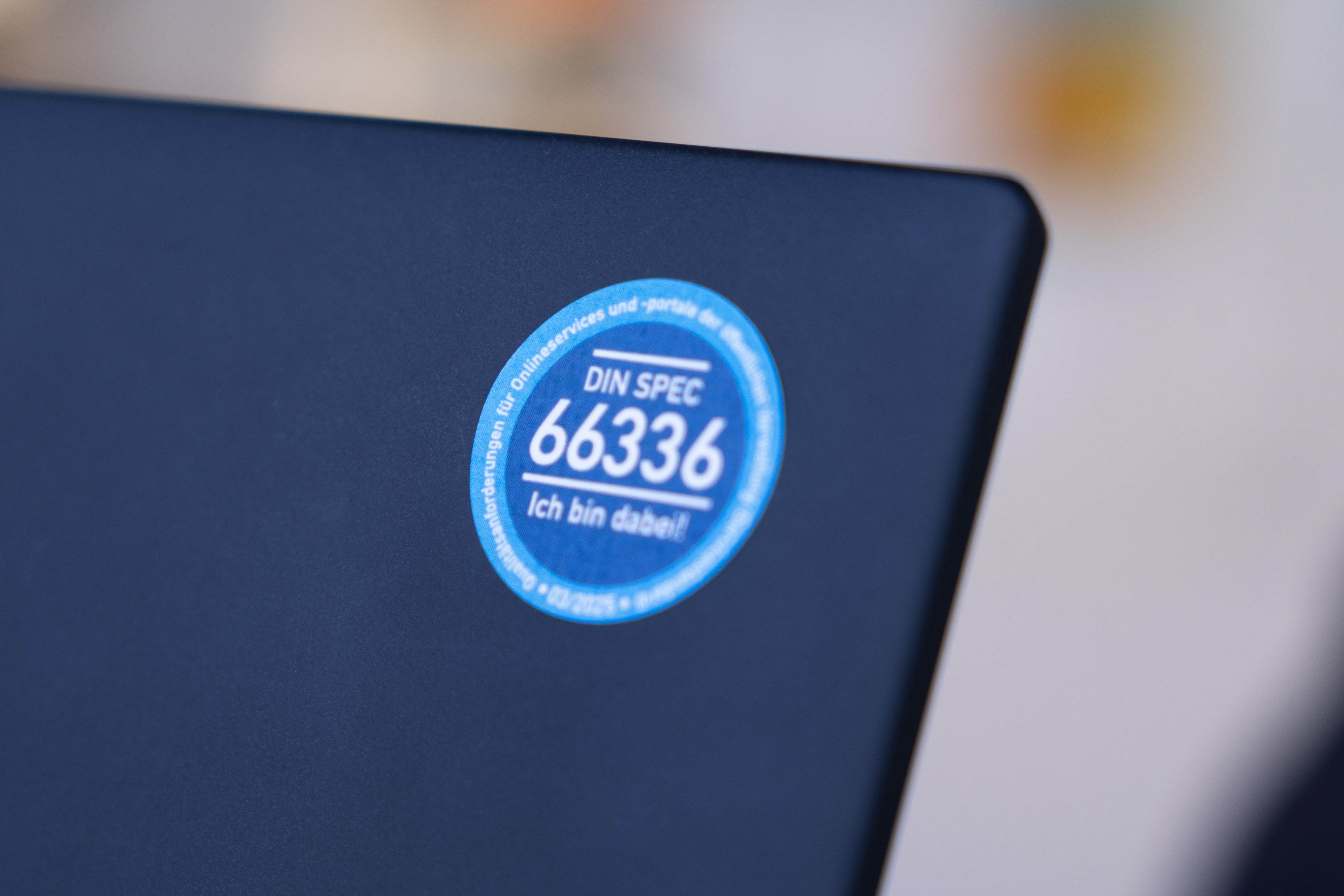 Ein blauer Sticker mit der Aufschrift „DIN SPEC 66336 – Ich bin dabei!” klebt auf der Rückseite eines dunklen Laptops. Der Hintergrund ist unscharf.