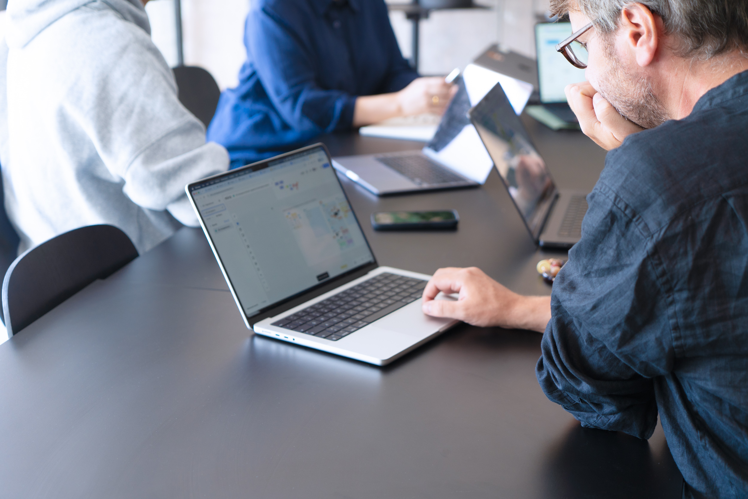 Eine Person bedient einen Laptop an einem Besprechungstisch, während im Hintergrund weitere Personen arbeiten.