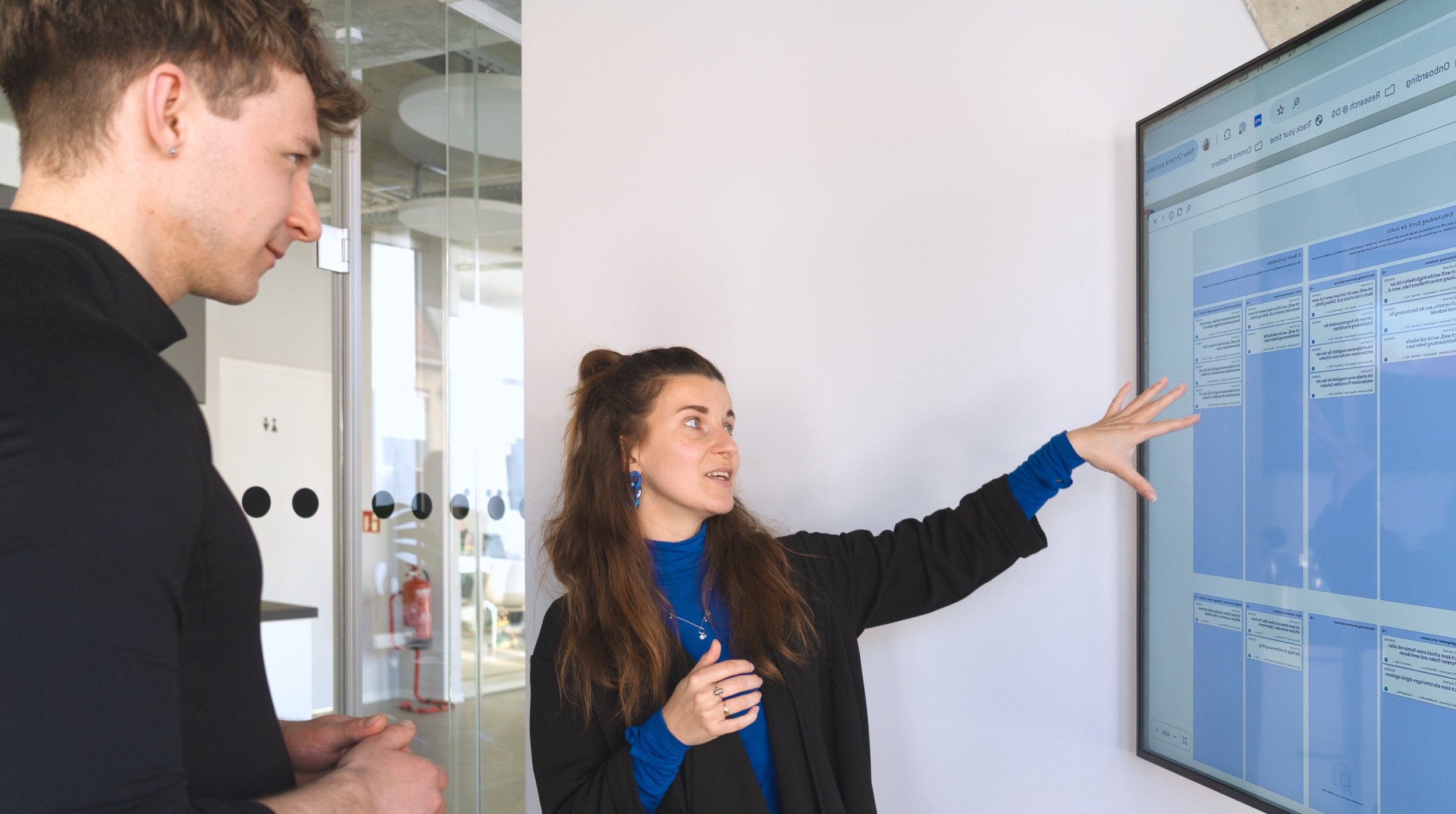 Zwei Personen stehen in einem Meetingraum vor einem großen Bildschirm. Die eine Person zeigt auf einen großen Bildschirm mit einem Task-Management-Board. Die andere steht daneben und hört zu.