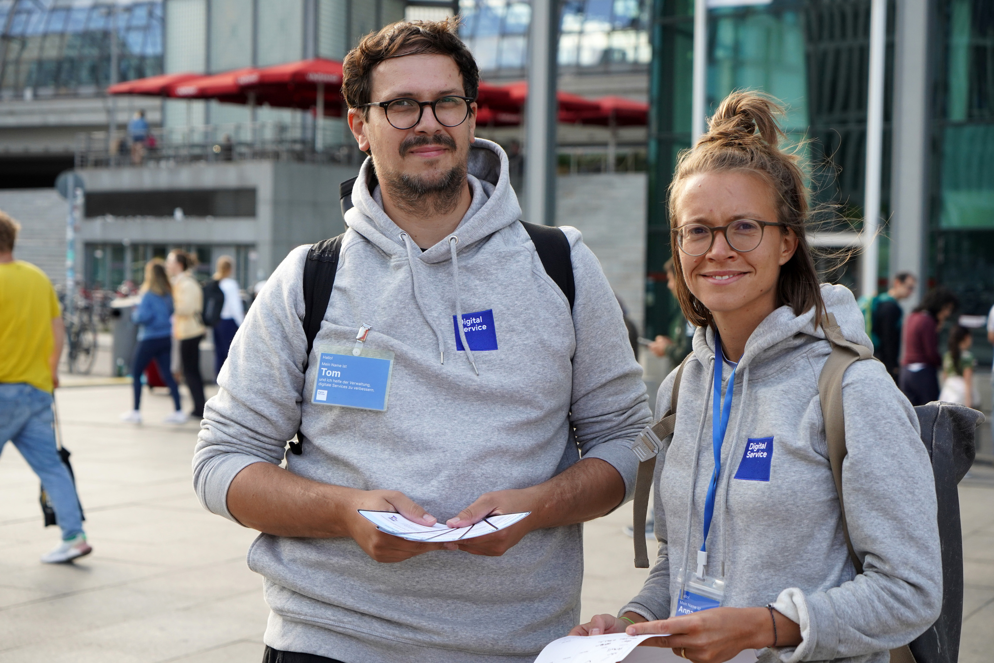 Ein jüngerer Mann und eine jüngere Frau stehen vor einem modernen Gebäude aus Glas und Beton, sie tragen beide die gleichen grauen Kaputzenpullover mit blauem Logo des DigitalService sowie blauen Namensschilder. Diese sagen Tom und Anna. Beide halten Zettel in ihren Händen. Diverse Menschen laufen hinter ihnen entlang.