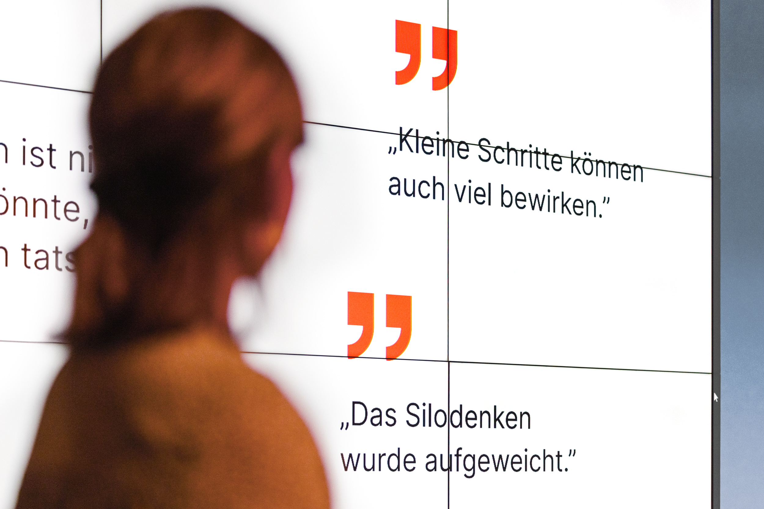 Eine Person steht vor einer großen digitalen Anzeige. Auf der Anzeige sind mehrere Textabschnitte und Zitate in deutscher Sprache zu sehen. Die Zitate lauten: „Kleine Schritte können auch viel bewirken“ und „Das Silodenken wurde aufgeweicht“.