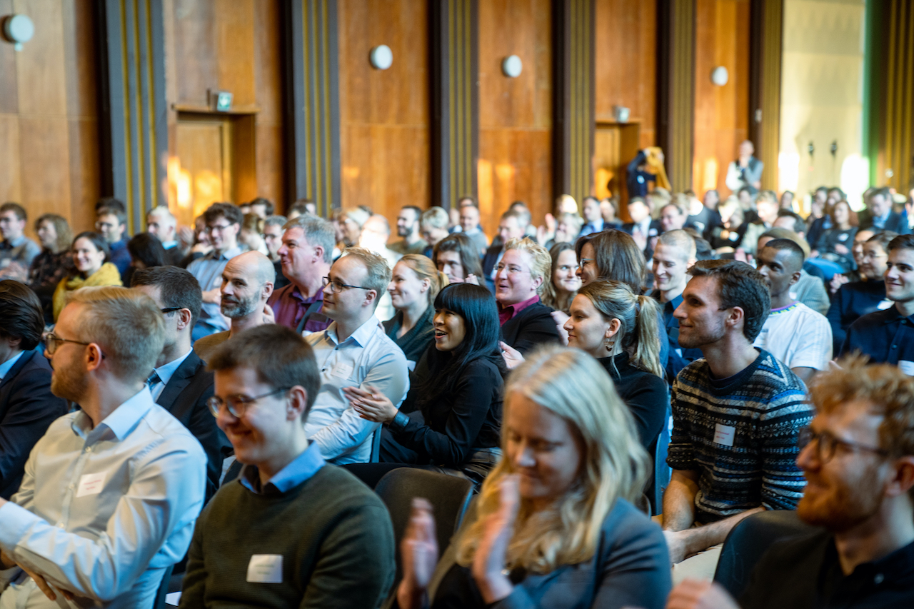 Abschlussveranstaltung Tech4Germany & Work4Germany. Viele Menschen sitzen in einem Raum. Im Hintergrund ist eine holzveräfelte Wand.