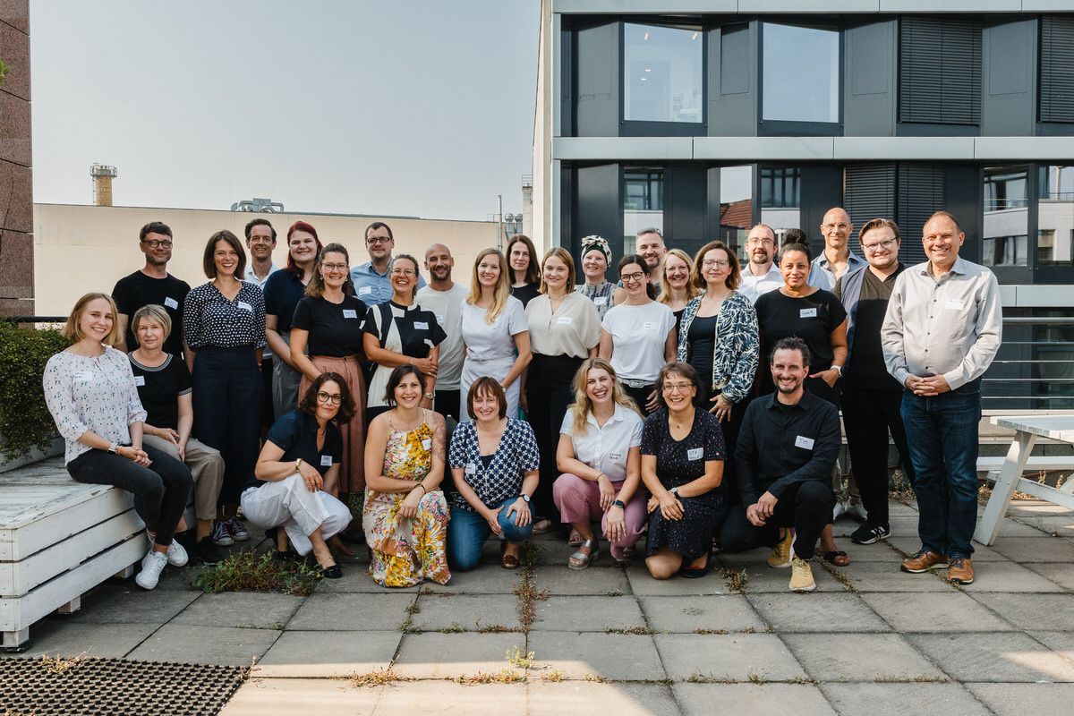 Eine Gruppe von Menschen, der 5. Jubiläumsjahrgang des Work4Germany Fellowships steht auf einer Terrasse aus Steinquadern. Links und rechts sind Bänke, im Hintergrund sind Bürogebäude zu sehen.