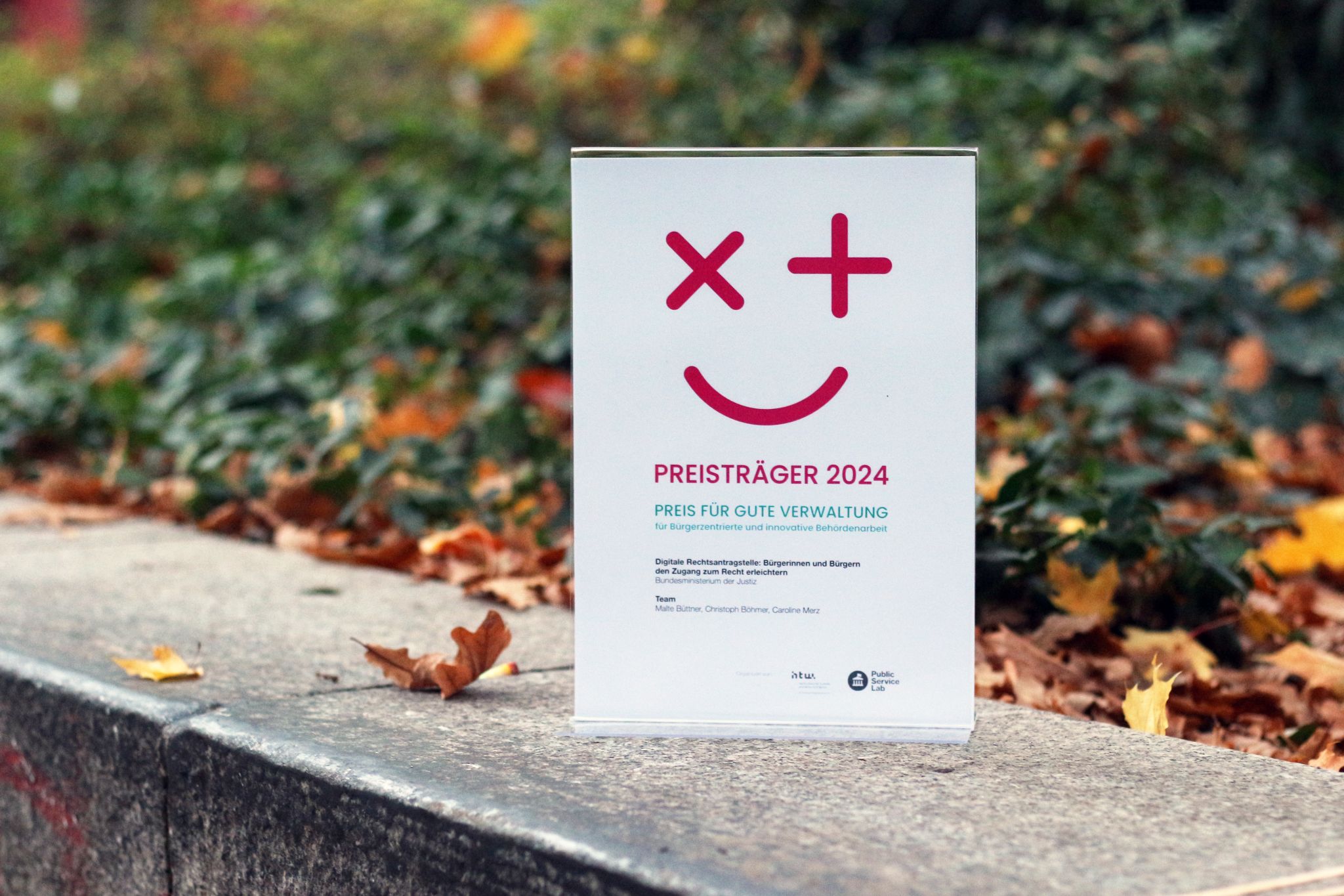 Eine Auszeichnung mit der Aufschrift „Preisträger 2024 – Preis für gute Verwaltung“ steht auf einem Steinpodest im Freien, umgeben von herbstlichem Laub.