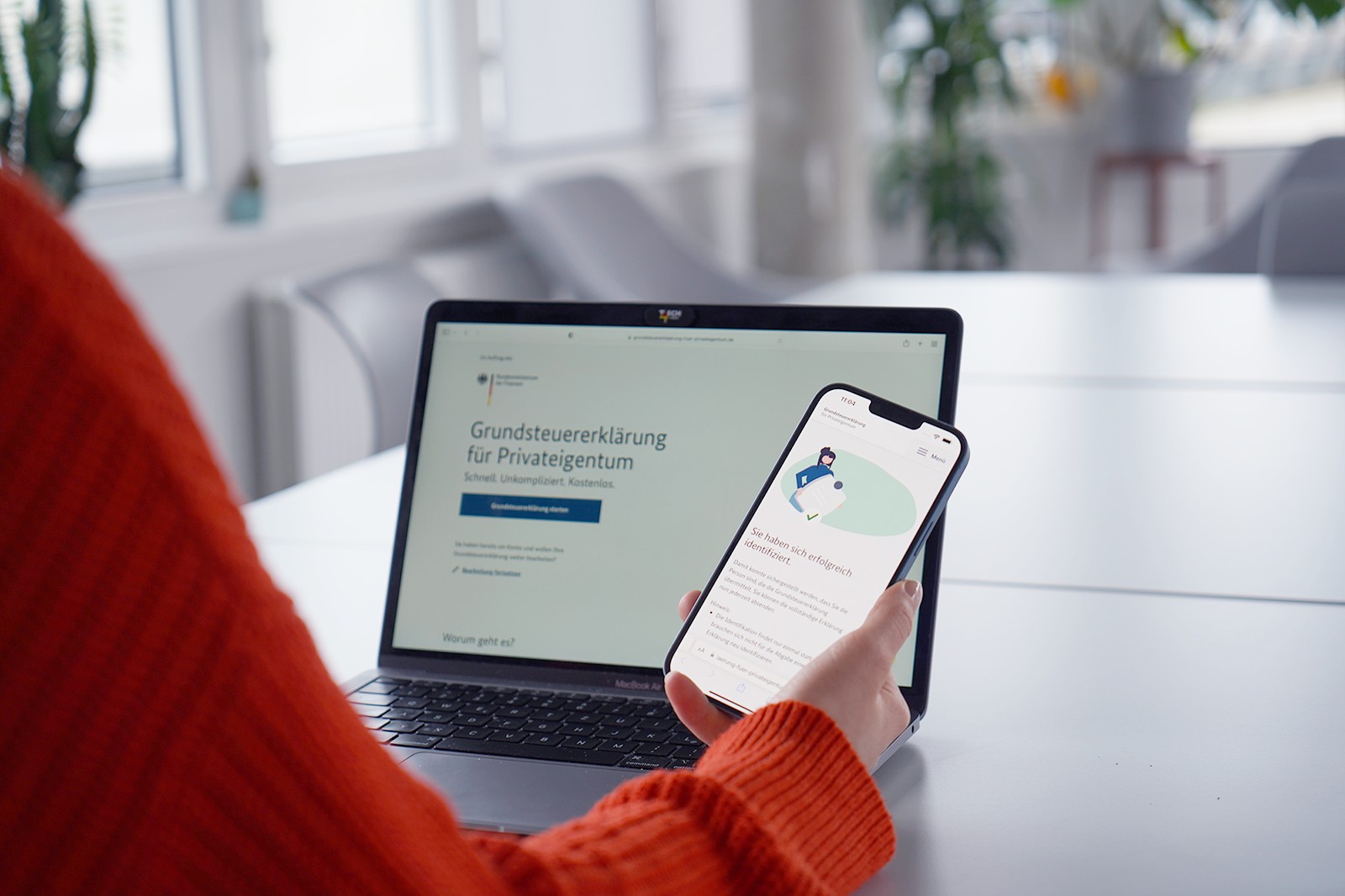 Eine Person in einem orangefarbenen Pullover hält ein Smartphone mit einer geöffneten Webseite, während im Hintergrund ein Laptop mit der Seite 